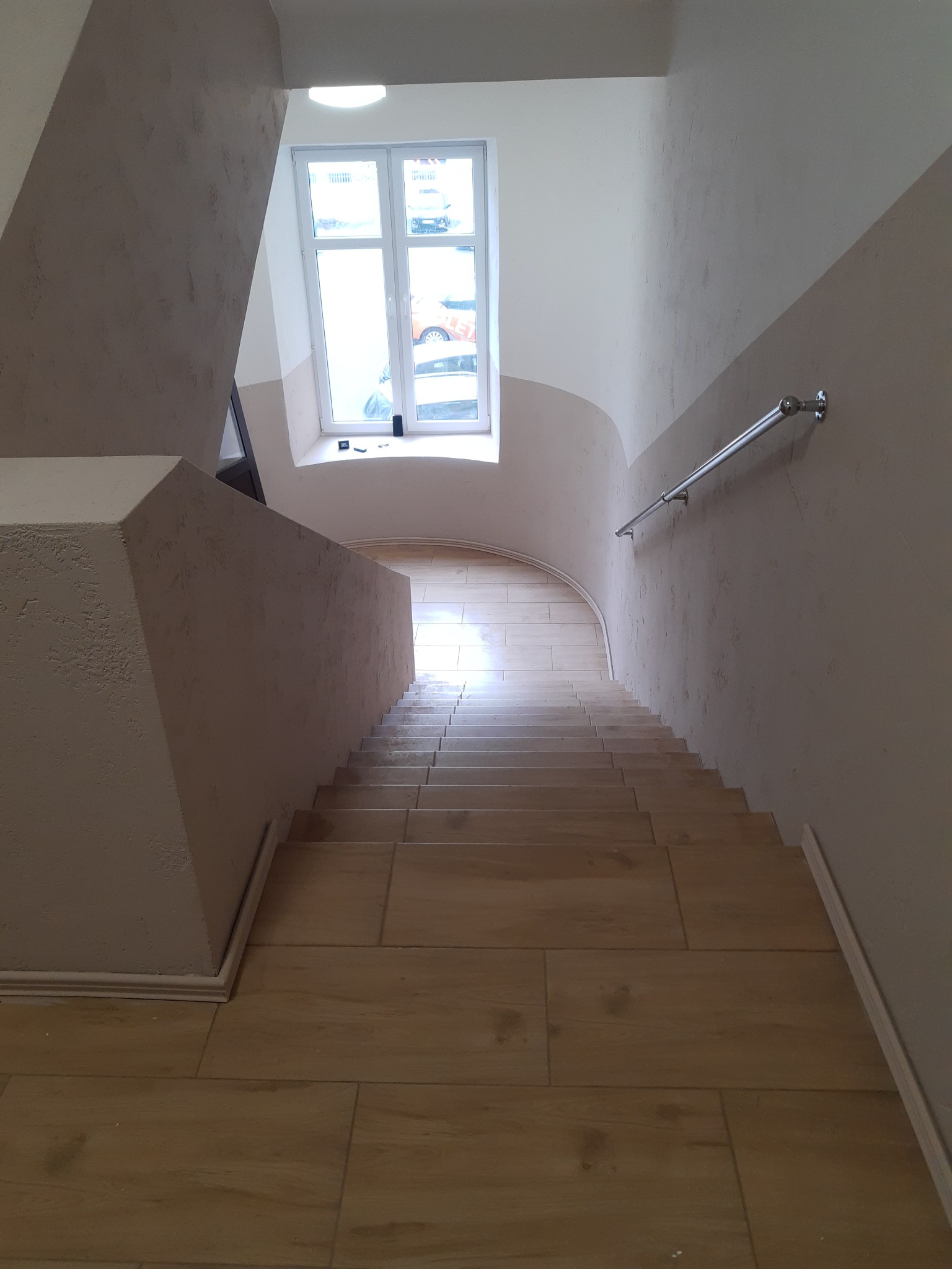 Wykończona klatka schodowa z ceramicznymi stopniami imitującymi drewno, ściany w odcieniach beżu z dekoracyjnym tynkiem, metalowa poręcz i okno z widokiem na ulicę.