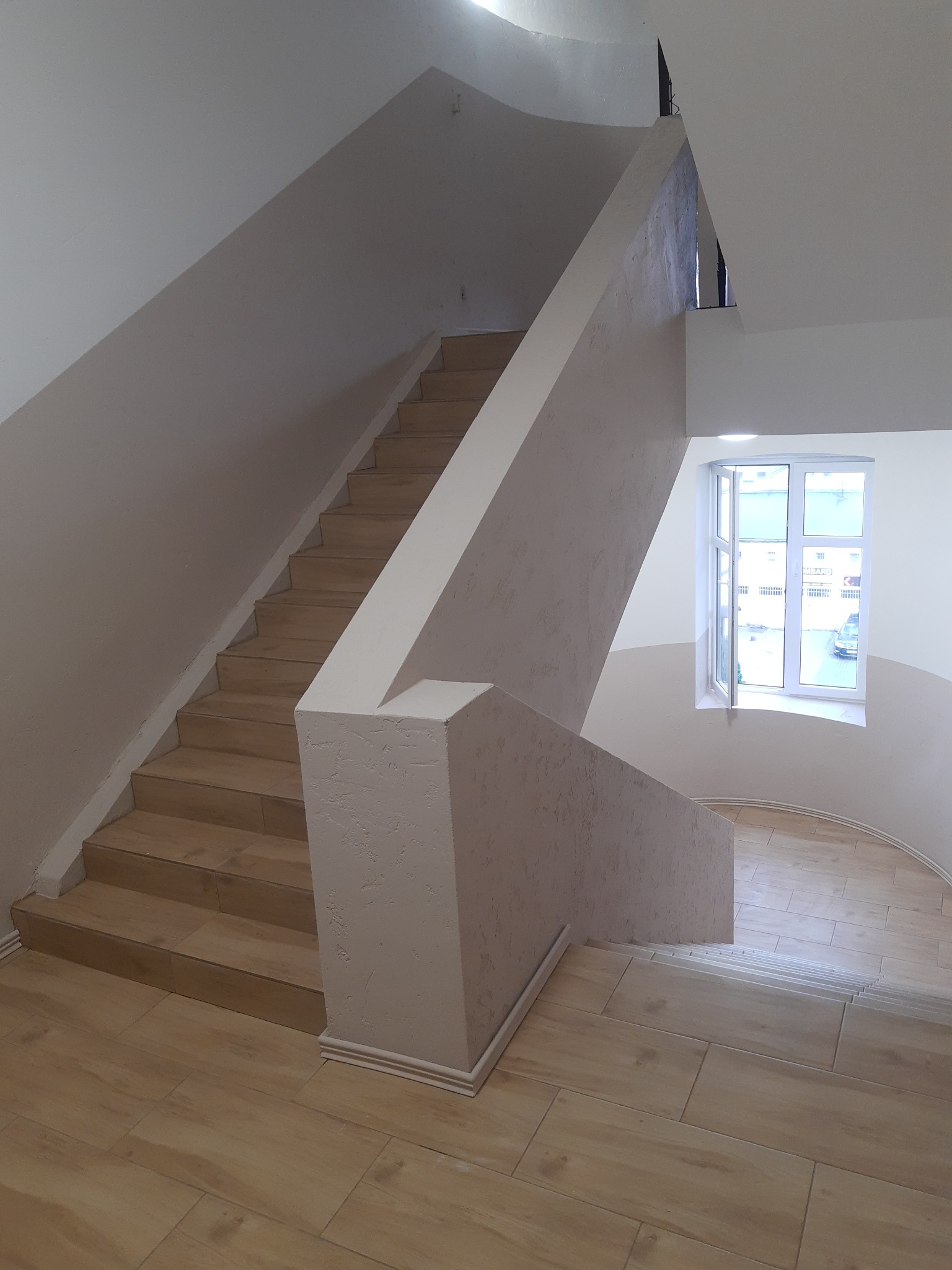 Klatka schodowa po remoncie z drewnopodobnymi płytkami na podłodze i schodach, ściany wykończone jasnym tynkiem strukturalnym, okno z widokiem na ulicę.