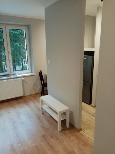 Jasne wnętrze mieszkania z drewnianą podłogą, białym regałem i widokiem na kuchnię z lodówką.