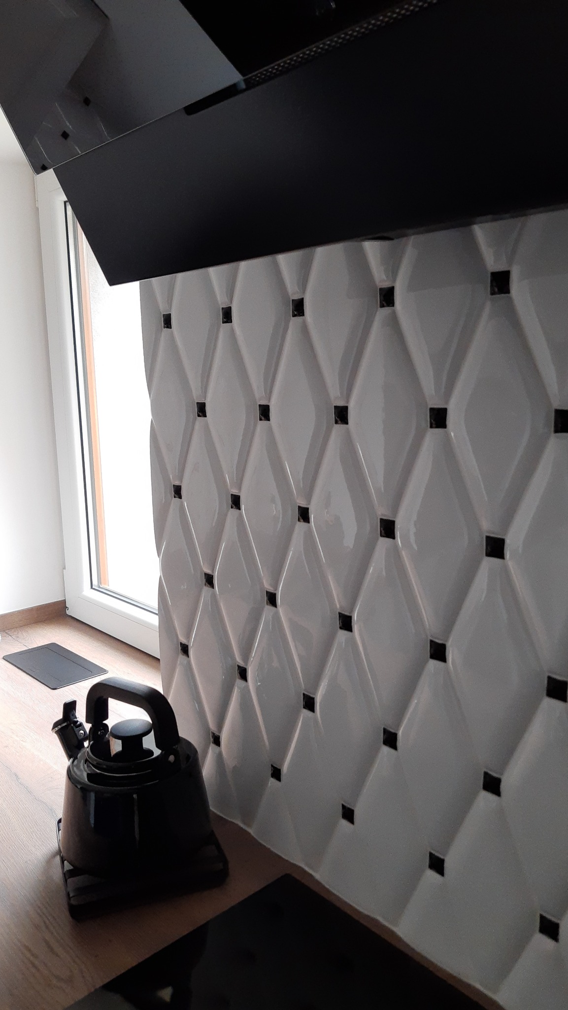Biała ściana z wypukłych, romboidalnych płytek z czarnymi akcentami, czarny czajnik na drewnianym blacie i fragment czarnego okapu kuchennego.