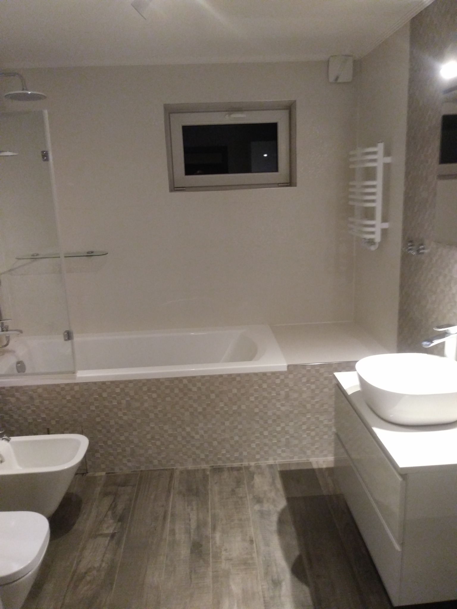 Nowoczesna łazienka z białą ceramiką, mozaikową obudową wanny i drewnopodobną podłogą, widok na kabinę prysznicową, umywalkę nablatową i grzejnik drabinkowy