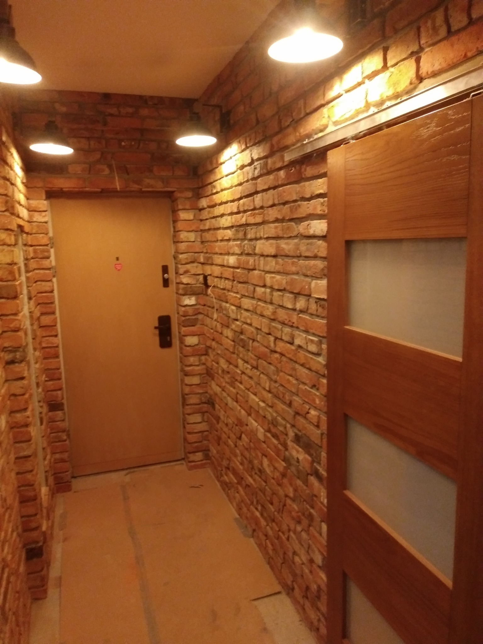 Korytarz z ceglanymi ścianami, oświetleniem punktowym i przesuwnymi drzwiami z drewnianymi ramami i matowym szkłem.