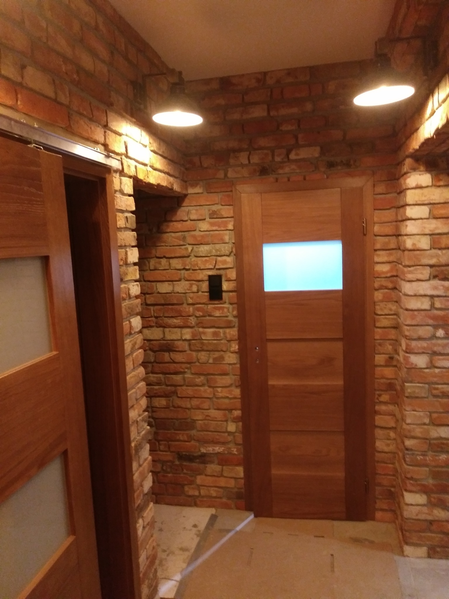 Wnętrze z wykończeniem ceglaną ścianą, oświetleniem punktowym i drewnianymi drzwiami z przeszkleniem, ukazujące połączenie surowego stylu z nowoczesnymi elementami.