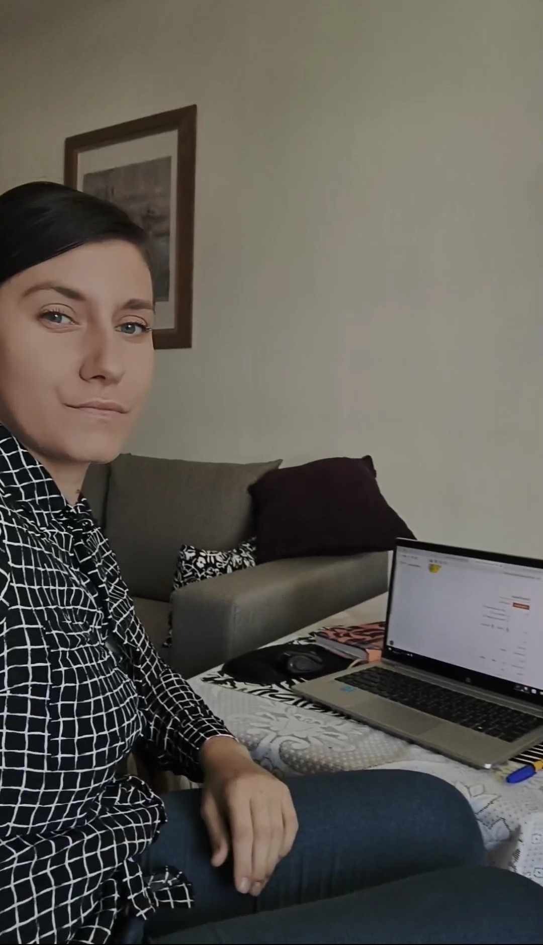 Kobieta w koszuli w kratę siedzi na kanapie z laptopem, prawdopodobnie podczas pracy zdalnej lub konsultacji online.