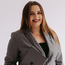 Beata Wójtowicz - Uśmiechnięta kobieta w szarym żakiecie trzymająca tablet Apple na białym tle, z widocznym naszyjnikiem z symbolami klucza i krzyża.