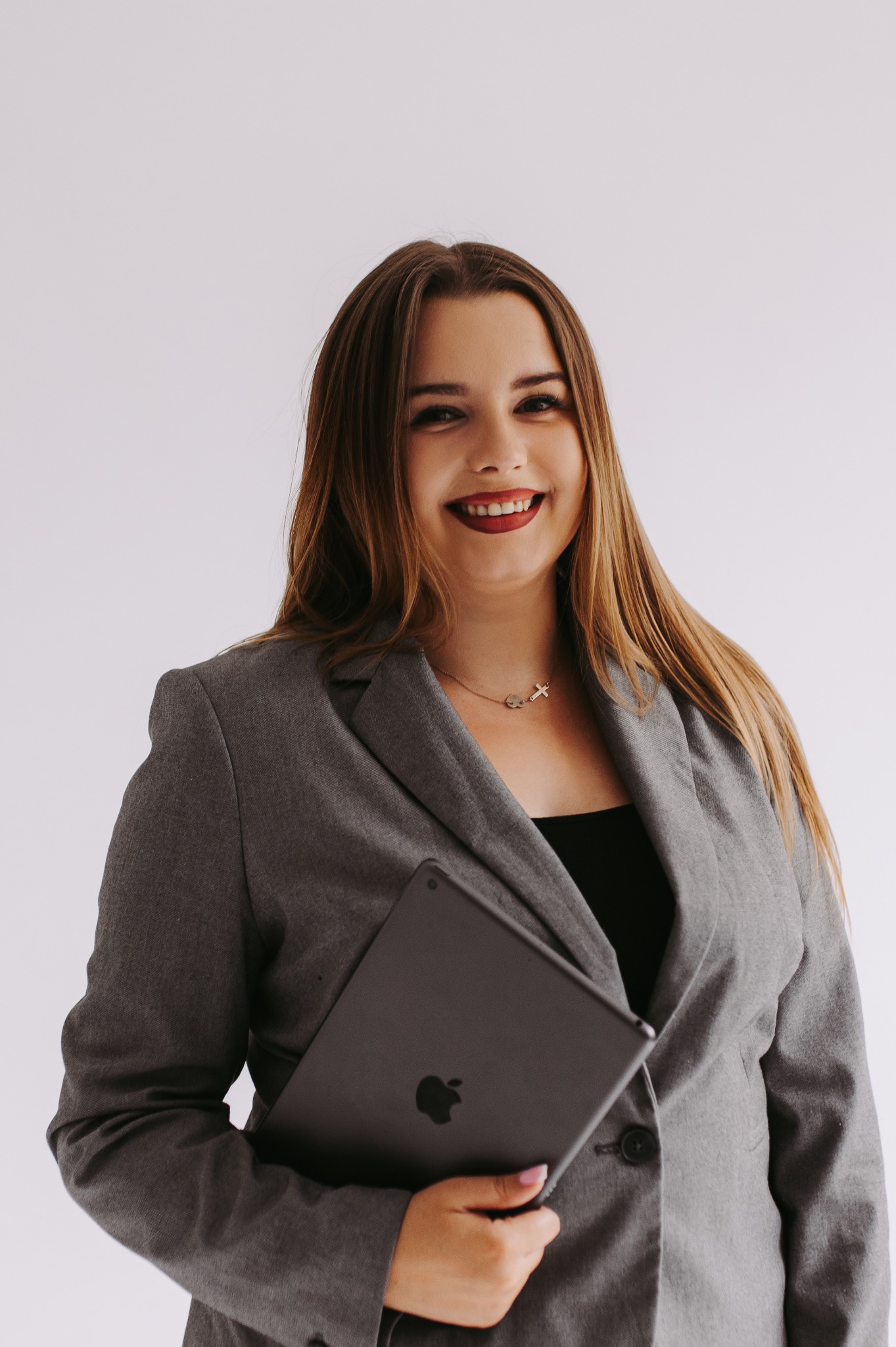 Uśmiechnięta kobieta w szarym żakiecie trzymająca tablet Apple na białym tle, z widocznym naszyjnikiem z symbolami klucza i krzyża.