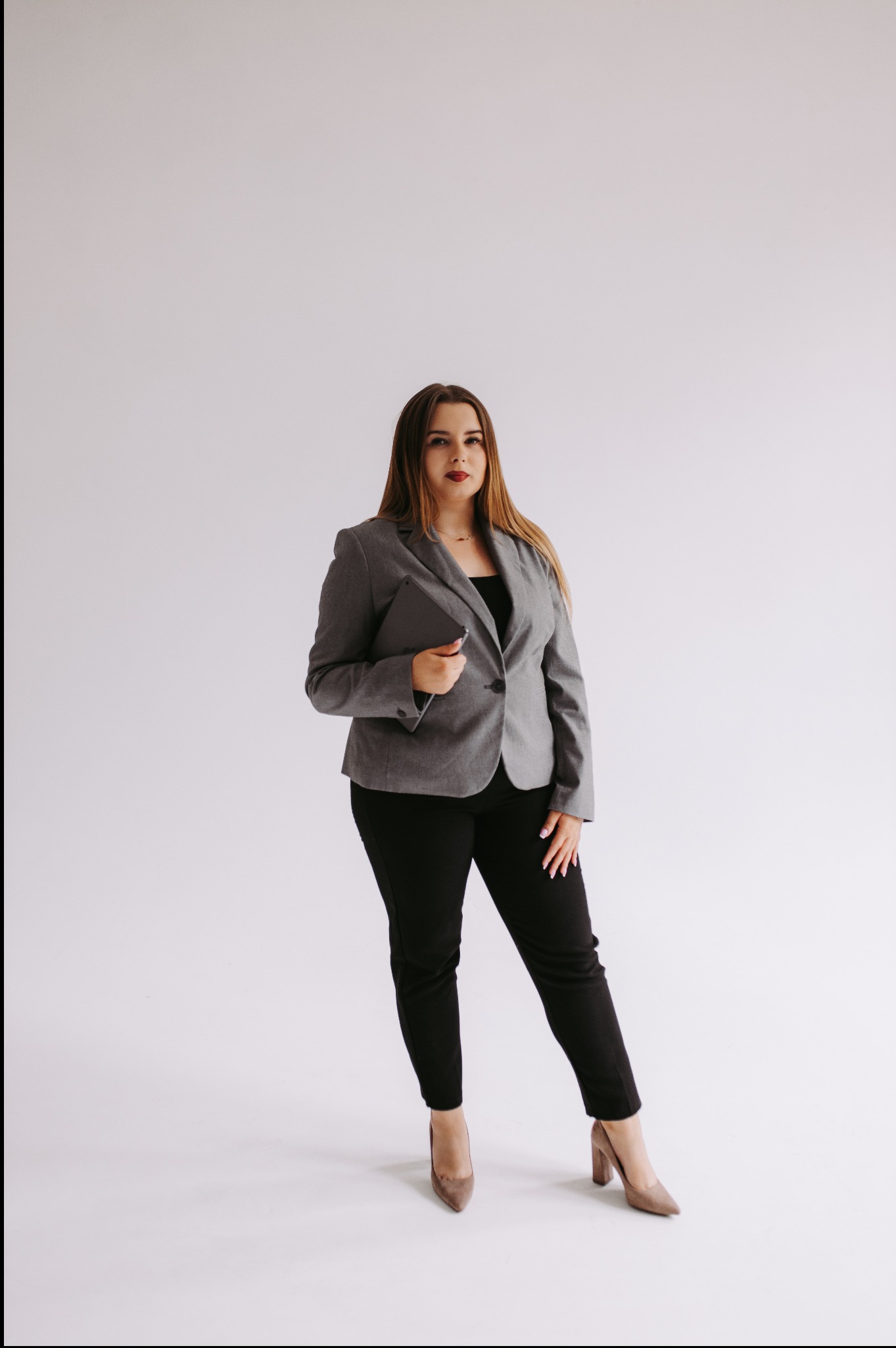 Kobieta w szarym żakiecie i czarnych spodniach, trzymająca tablet na białym tle, prezentująca profesjonalny wizerunek.
