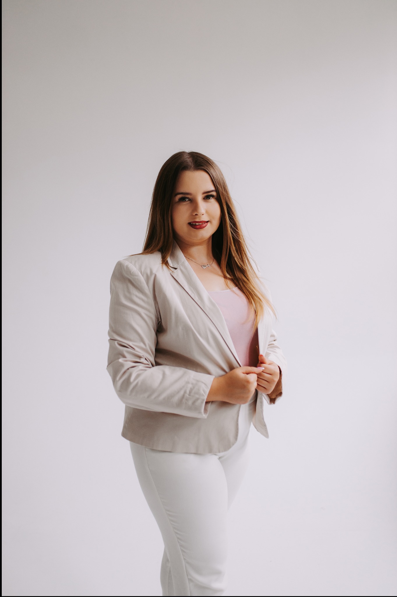Profesjonalny portret kobiety w beżowym żakiecie i białych spodniach na jasnym tle, z delikatnym uśmiechem i subtelną biżuterią, sugerujący reprezentanta firmy ubezpieczeniowej.