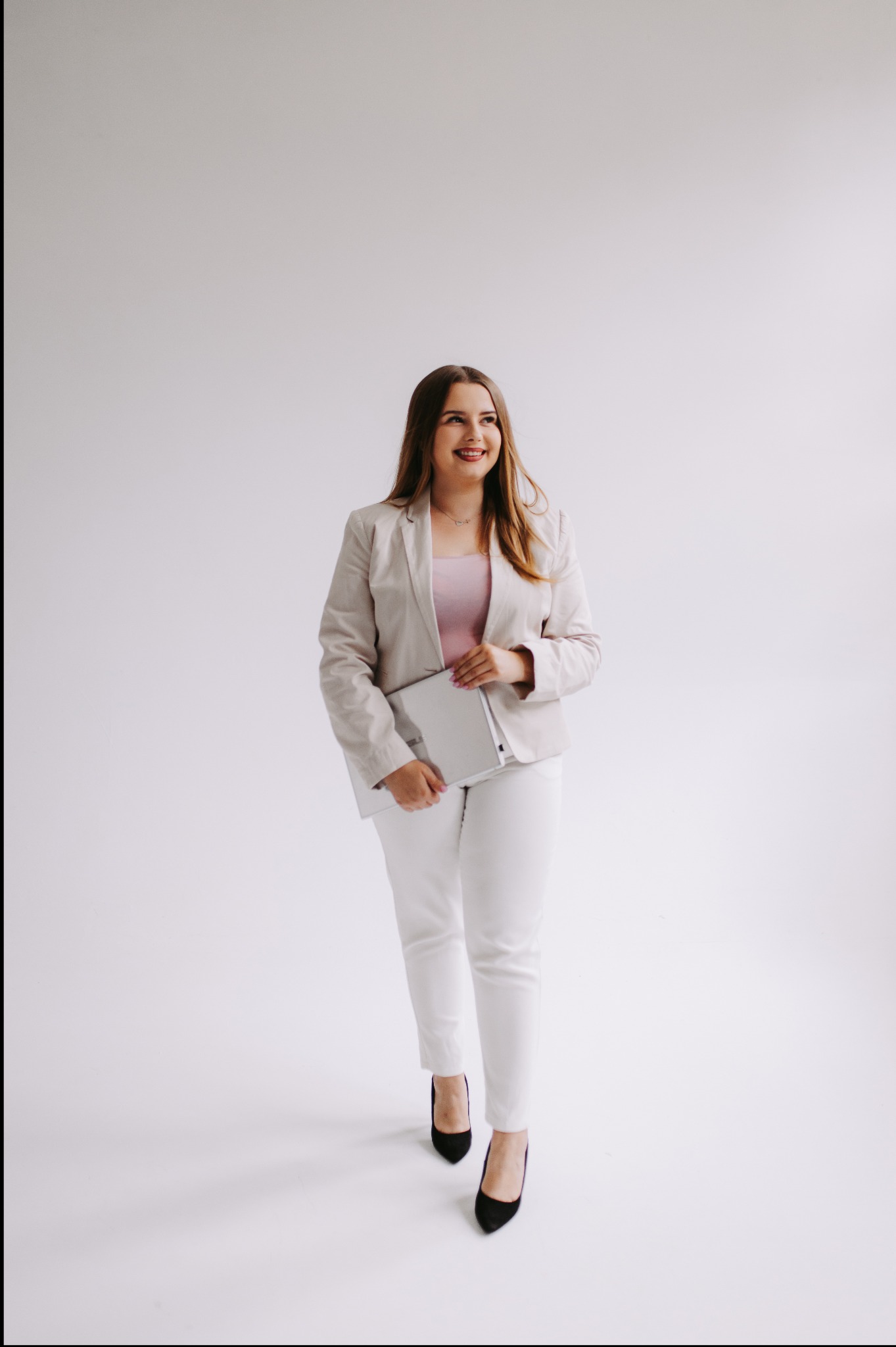 Uśmiechnięta kobieta w jasnym garniturze i czarnych szpilkach, trzymająca srebrny laptop na białym tle, patrzy w bok.