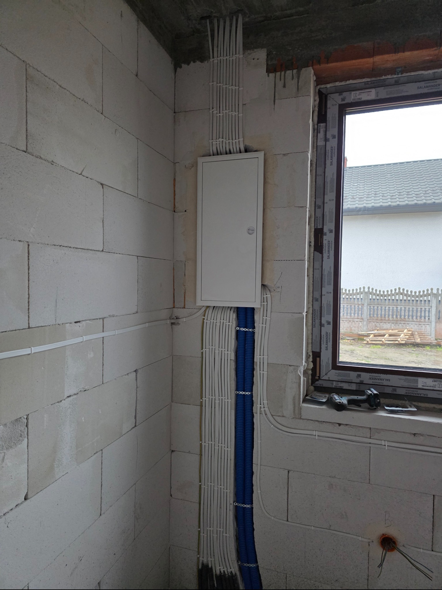 Biała skrzynka elektryczna na surowej ścianie z białych bloczków, z widocznymi białymi i niebieskimi rurkami instalacyjnymi. W tle okno z widokiem na zewnątrz.