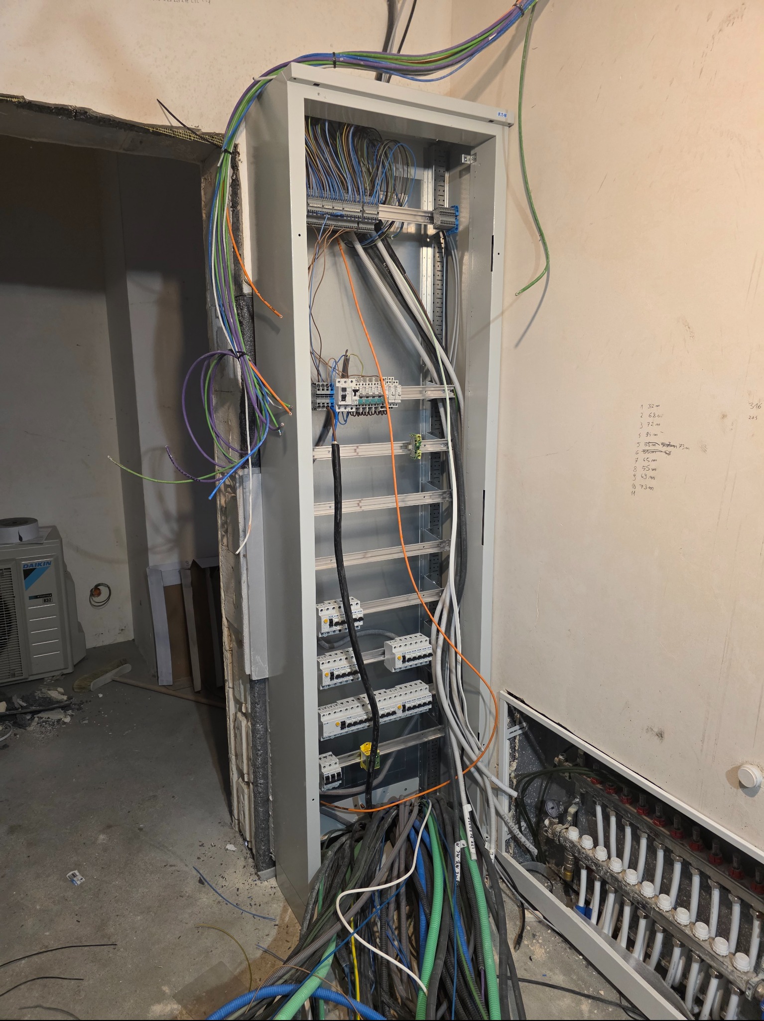 Otwarta szafa rozdzielcza z licznymi kablami w różnych kolorach, widoczne bezpieczniki i listwy zaciskowe. Na ścianie notatki z pomiarami. W tle klimatyzator.