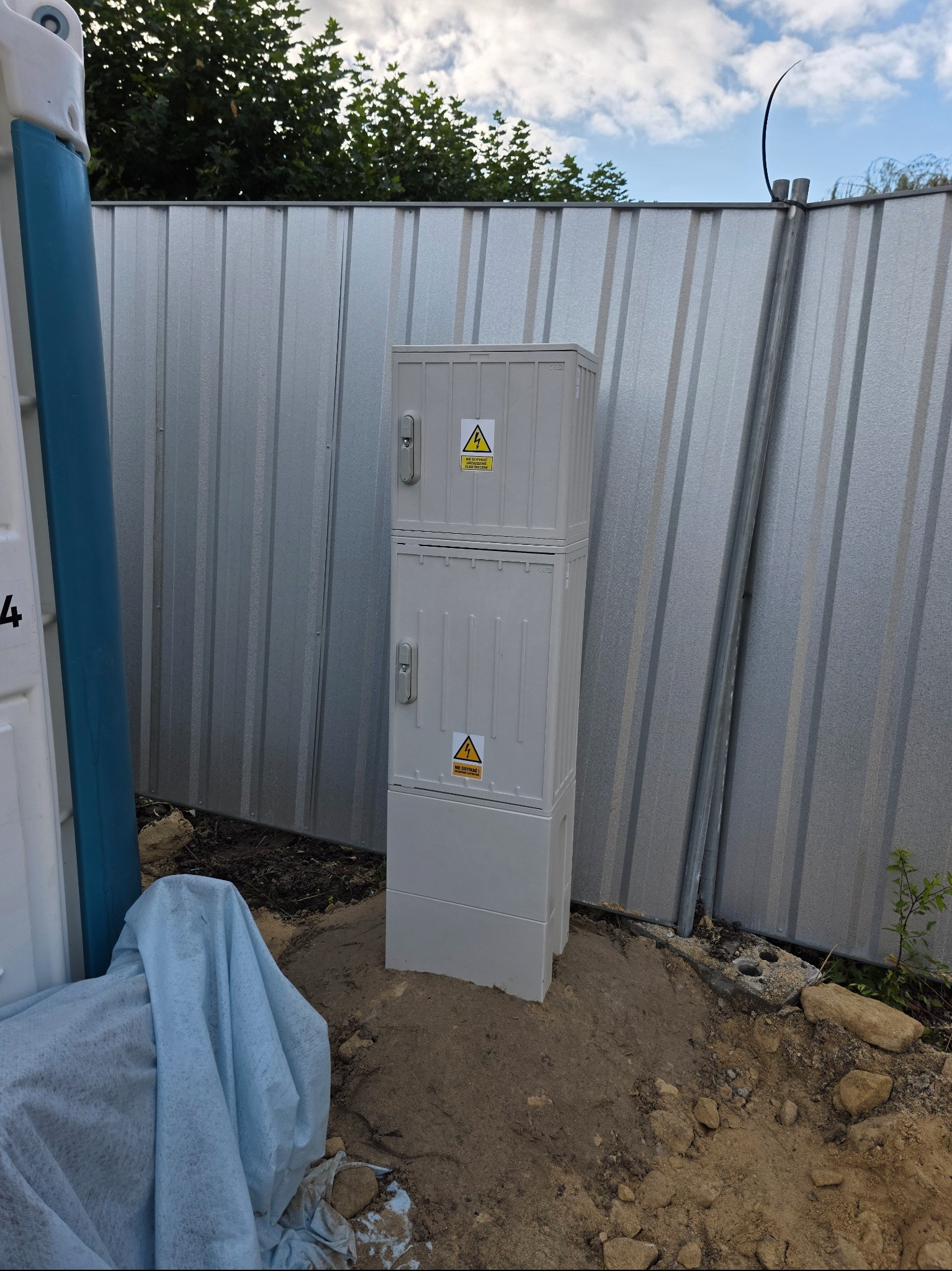 Zmontowana, modułowa skrzynka elektryczna z ostrzeżeniami, umieszczona na ziemi przy metalowym ogrodzeniu. Widoczne elementy instalacji i otoczenie placu budowy.