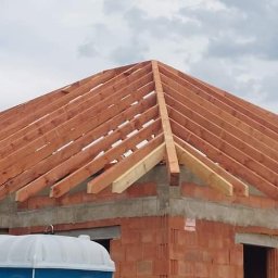 Czaro-dachy - Wiązar dachowy z drewna na murowanym budynku w trakcie budowy, widoczne niebo i drzewa w tle, na pierwszym planie biały przenośny WC.