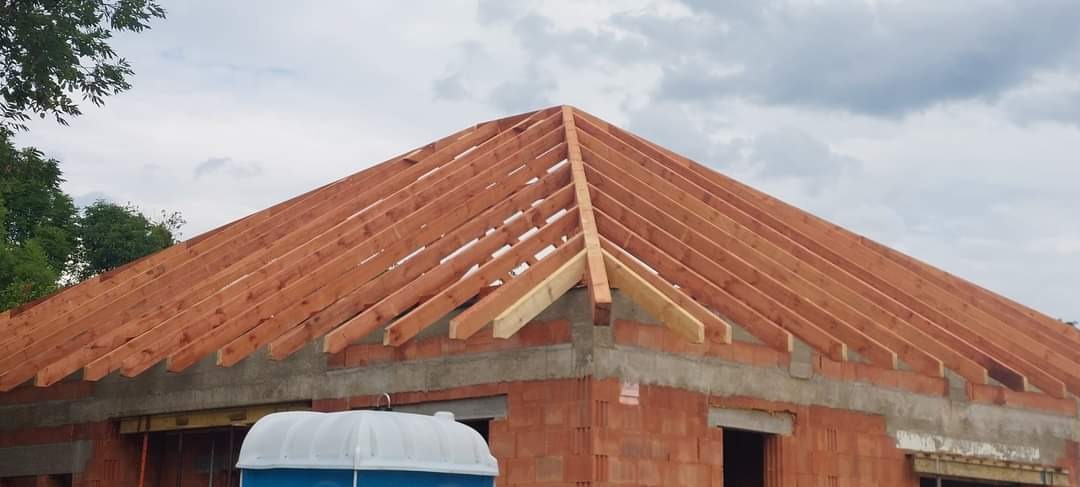 Wiązar dachowy z drewna na murowanym budynku w trakcie budowy, widoczne niebo i drzewa w tle, na pierwszym planie biały przenośny WC.