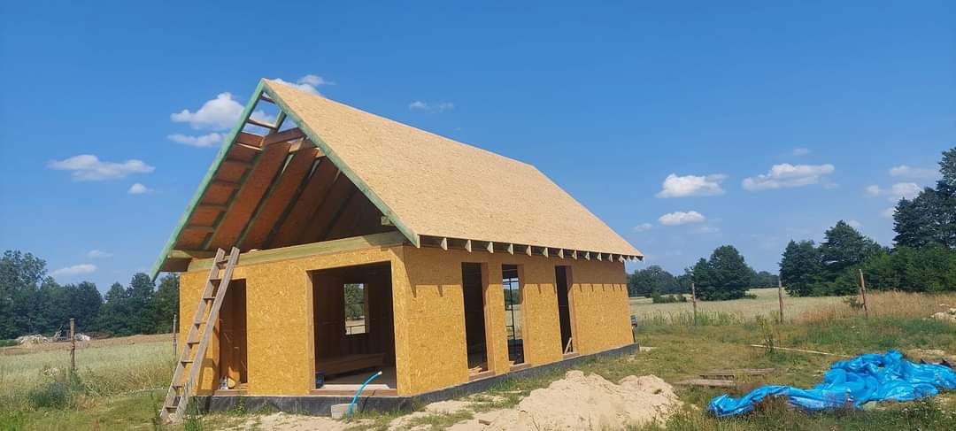 Budowa domu w stanie surowym otwartym: widoczna konstrukcja ścian z płyt OSB i więźba dachowa z deskowaniem, drabina oparta o ścianę szczytową, błękitne niebo w tle.