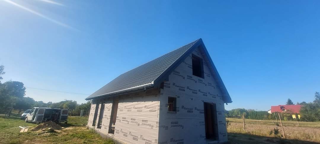 Dom w trakcie budowy z zamontowanym dachem z blachy w kolorze grafitowym i orynnowaniem, ściany zabezpieczone membraną, widoczny samochód dostawczy na posesji.