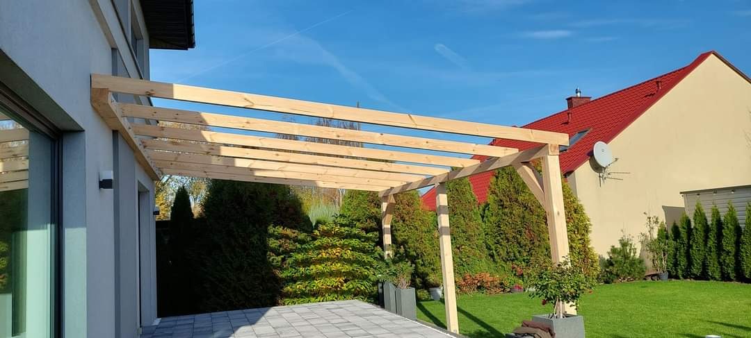 Drewniana konstrukcja pergoli w ogrodzie, widoczne elementy więźby dachowej, tło stanowi dom z czerwonym dachem i zielona roślinność.