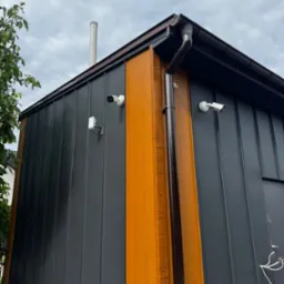 Nowoczesny budynek z szarej blachy trapezowej, z elementami dekoracyjnymi z drewna i dwiema kamerami monitoringu na elewacji. Widok z dołu na róg budynku.