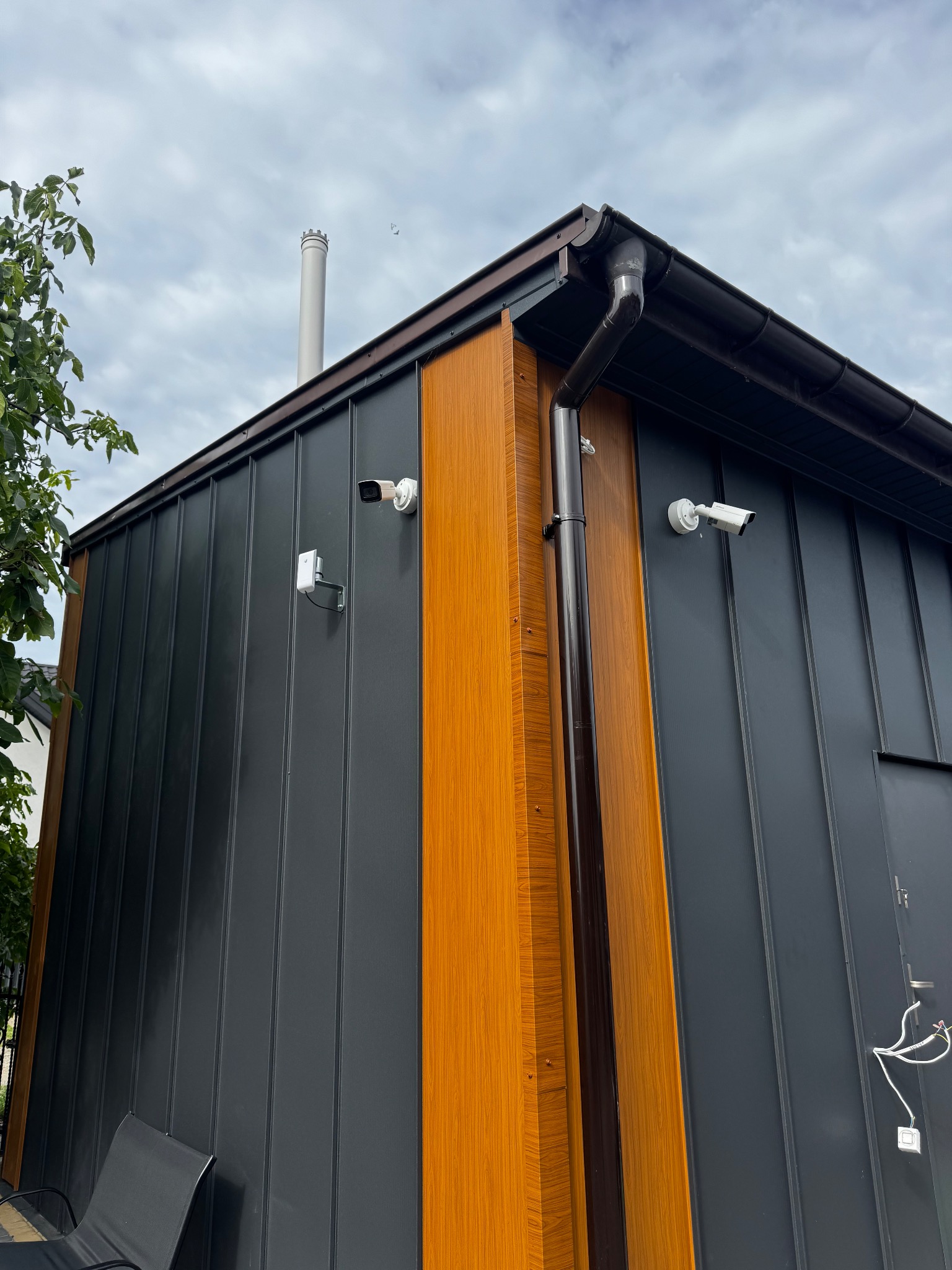 Nowoczesny budynek z szarej blachy trapezowej, z elementami dekoracyjnymi z drewna i dwiema kamerami monitoringu na elewacji. Widok z dołu na róg budynku.