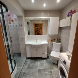 Taras I kompania - Wyremontowana łazienka w szaro-białej kolorystyce: kabina prysznicowa z czarnymi profilami, umywalka z szafką, toaleta, pralka Bosch, szare płytki imitujące marmur na podłodze i ścianach, lustro...