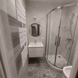 Taras I kompania - Wykończona łazienka z kabiną prysznicową, umywalką z szafką, grzejnikiem drabinkowym i wzorzystą podłogą; odbicie osoby robiącej zdjęcie w okrągłym lustrze.