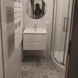 Nowoczesna aranżacja łazienki w Piasecznie: biała szafka pod umywalką, okrągłe lustro z widocznym odbiciem osoby, grzejnik drabinkowy oraz kabina prysznicowa z zaokrąglonymi drzwiami, całość...