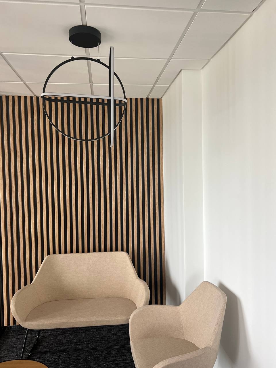 Nowoczesne wykończenie wnętrza w Piasecznie: drewniana ściana z pionowych listew, designerska lampa wisząca i beżowe fotele tworzą minimalistyczną przestrzeń.