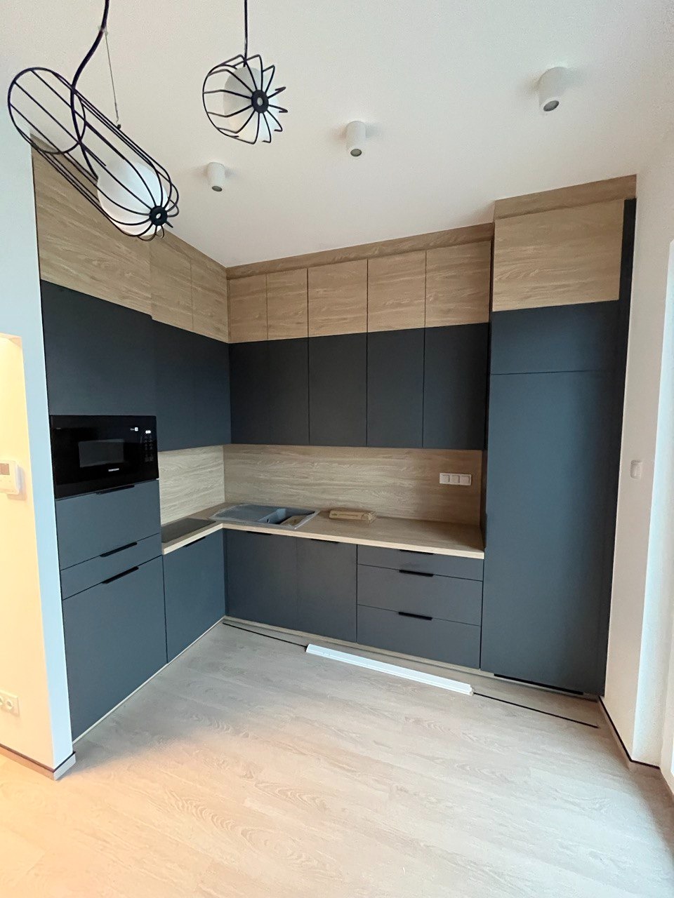 Nowoczesna kuchnia w odcieniach szarości i drewna z designerskimi lampami wiszącymi, minimalistyczne fronty szafek bez uchwytów, jasna drewniana podłoga.