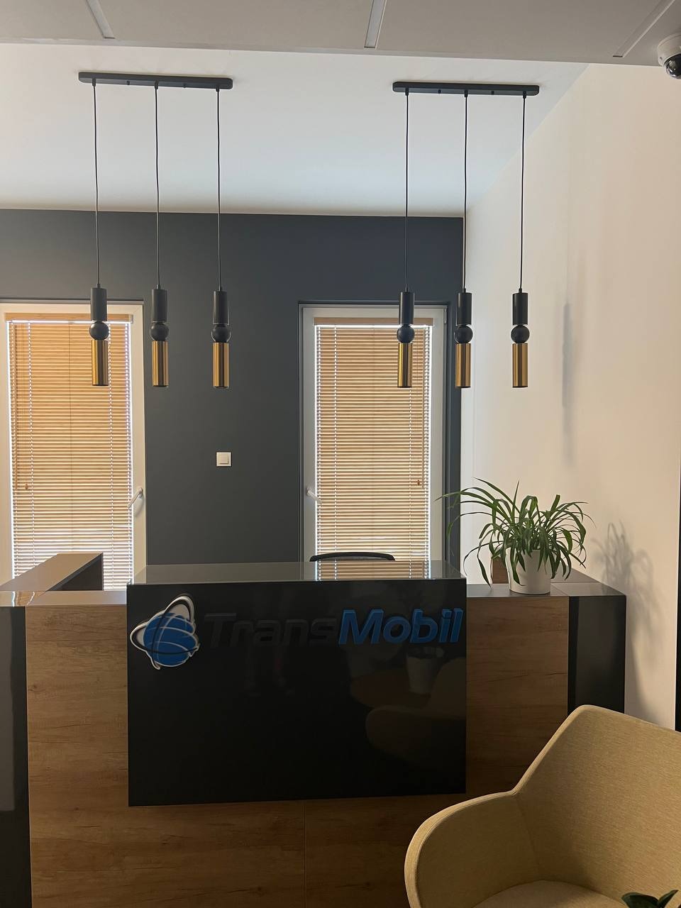 Recepcja biurowa z drewnianym wykończeniem, czarnym blatem z logo 'TransMobil', oświetlona trzema wiszącymi lampami z elementami złotymi i czarnymi, roślina doniczkowa na blacie, widok na okna...