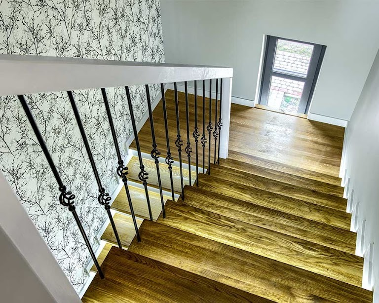 Schody drewniane z czarnymi, metalowymi balustradami i białymi poręczami, wykończone tapetą w delikatny wzór gałązek na ścianie, z widocznym oknem na podeście.