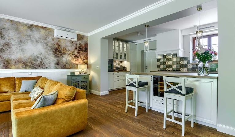 Salon z aneksem kuchennym w stylu eklektycznym: złoty narożnik, ściana z wzorzystą tapetą, drewniana podłoga, białe krzesła barowe przy wyspie kuchennej z dekoracyjną kratką z płytek.
