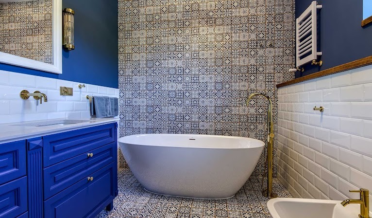 Elegancka łazienka z wolnostojącą wanną, granatowymi szafkami umywalkowymi, złotą armaturą i dekoracyjnymi płytkami na ścianach oraz podłodze.