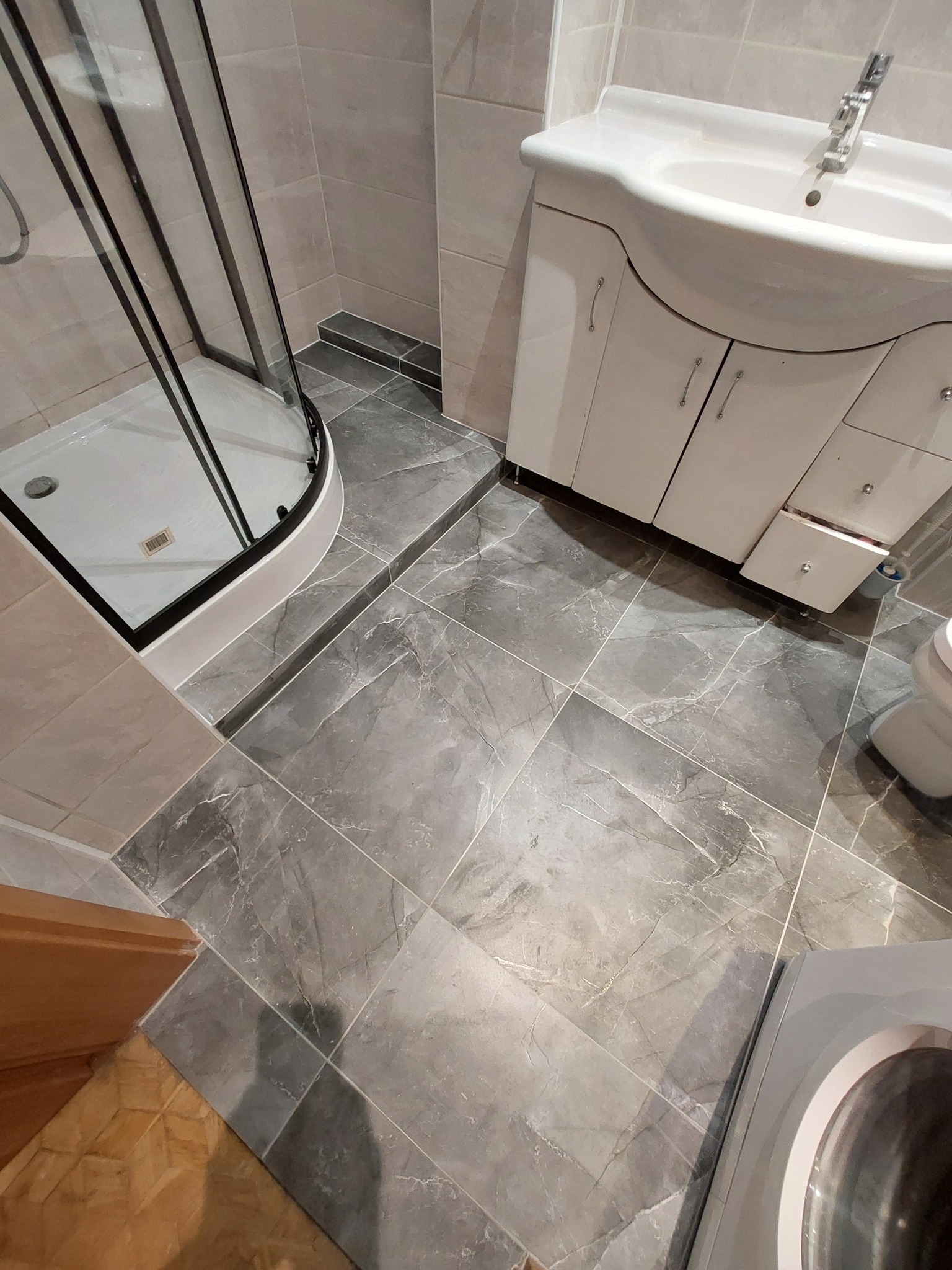 Wykończona łazienka z szarą marmurową podłogą, kabiną prysznicową z czarnymi ramami, umywalką z szafką i częściowo widoczną pralką.