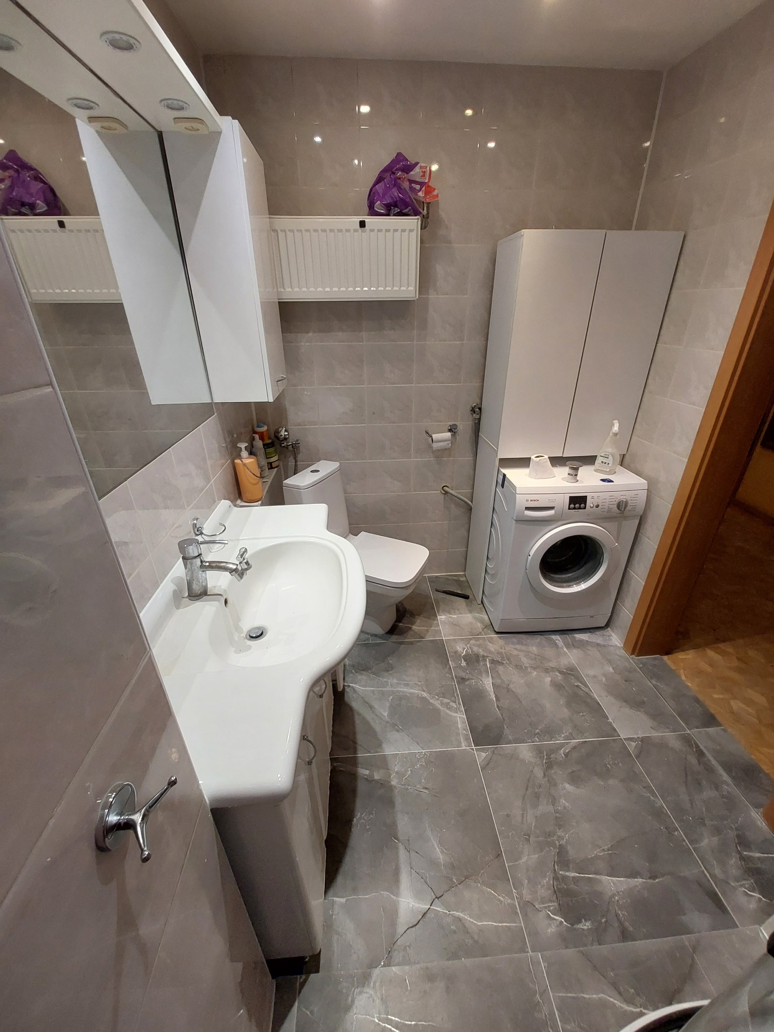 Wykończona łazienka z umywalką na szafce, toaletą, pralką i szafką wiszącą, wyłożona płytkami imitującymi marmur w odcieniach szarości.