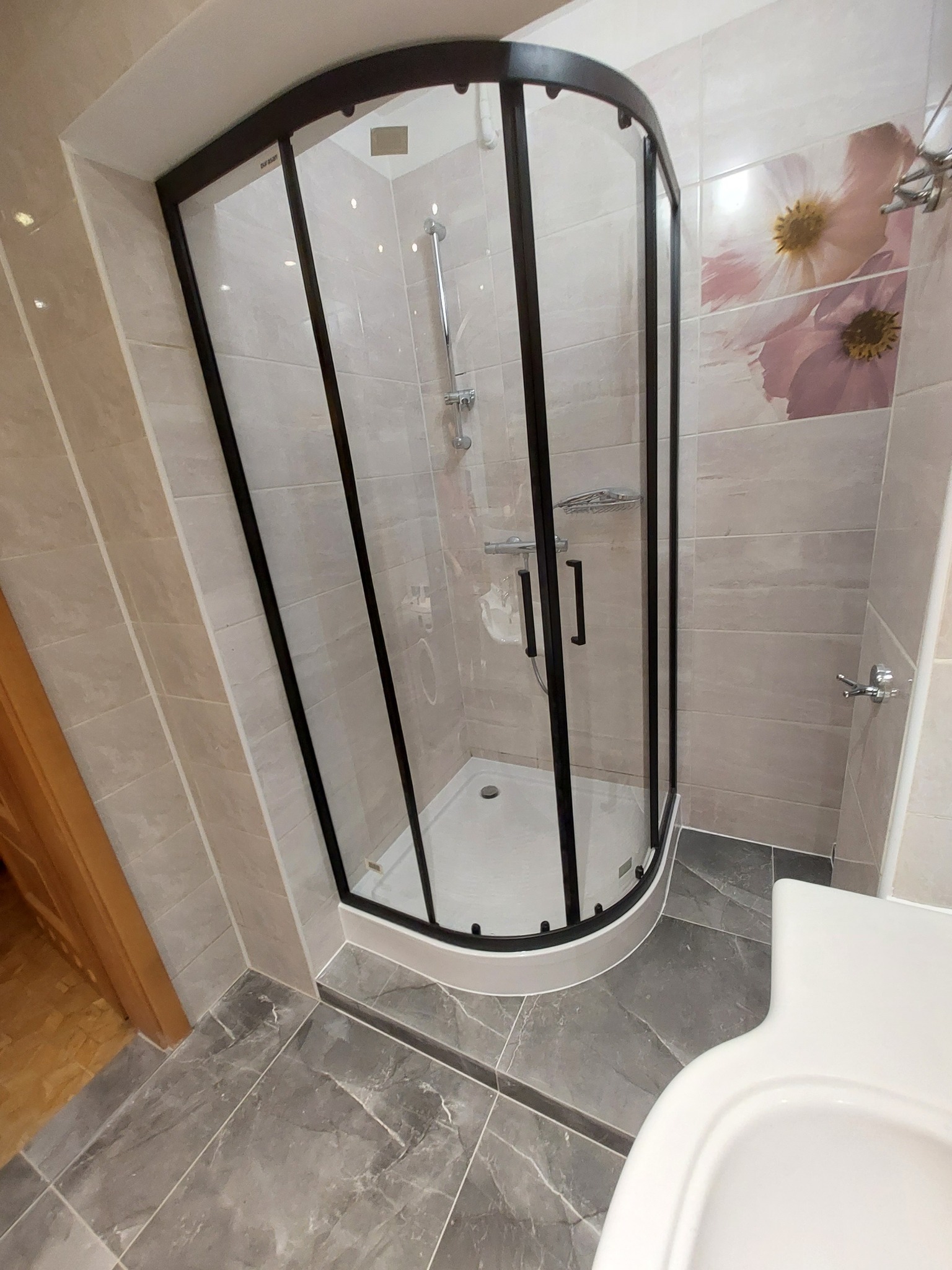 Nowoczesna kabina prysznicowa z czarną ramą w narożnej łazience wykończonej płytkami w odcieniach szarości i beżu, z dekoracyjnym motywem kwiatowym na ścianie i widoczną umywalką.