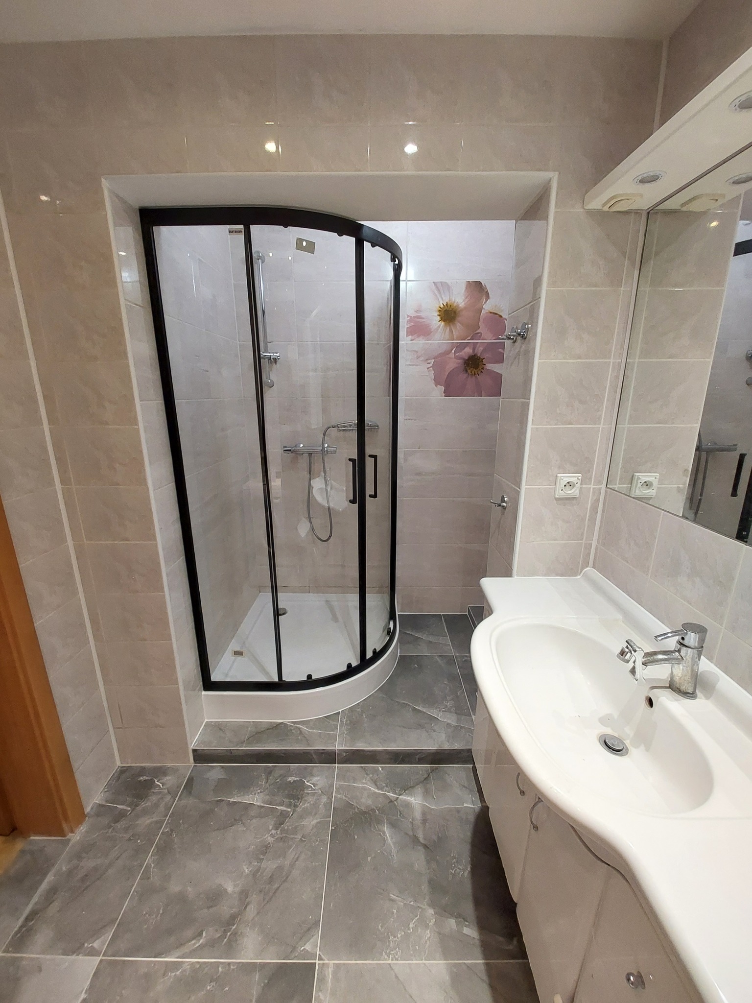 Wykończona łazienka z kabiną prysznicową z czarną ramą, umywalką w zabudowie i szarą marmurową podłogą, akcent w postaci kwiatowego motywu na ścianie prysznica.