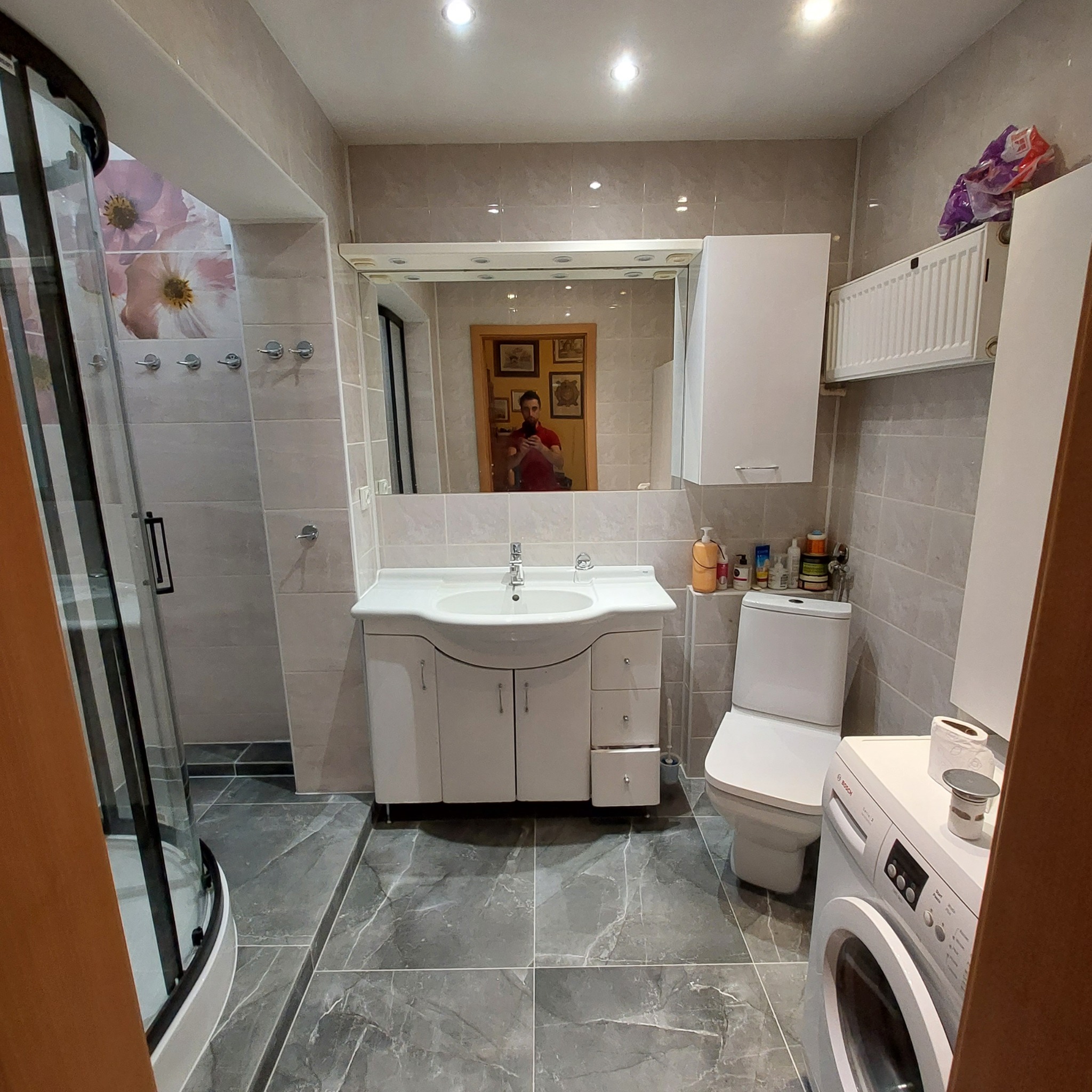 Wyremontowana łazienka w szaro-białej kolorystyce: kabina prysznicowa z czarnymi profilami, umywalka z szafką, toaleta, pralka Bosch, szare płytki imitujące marmur na podłodze i ścianach, lustro...