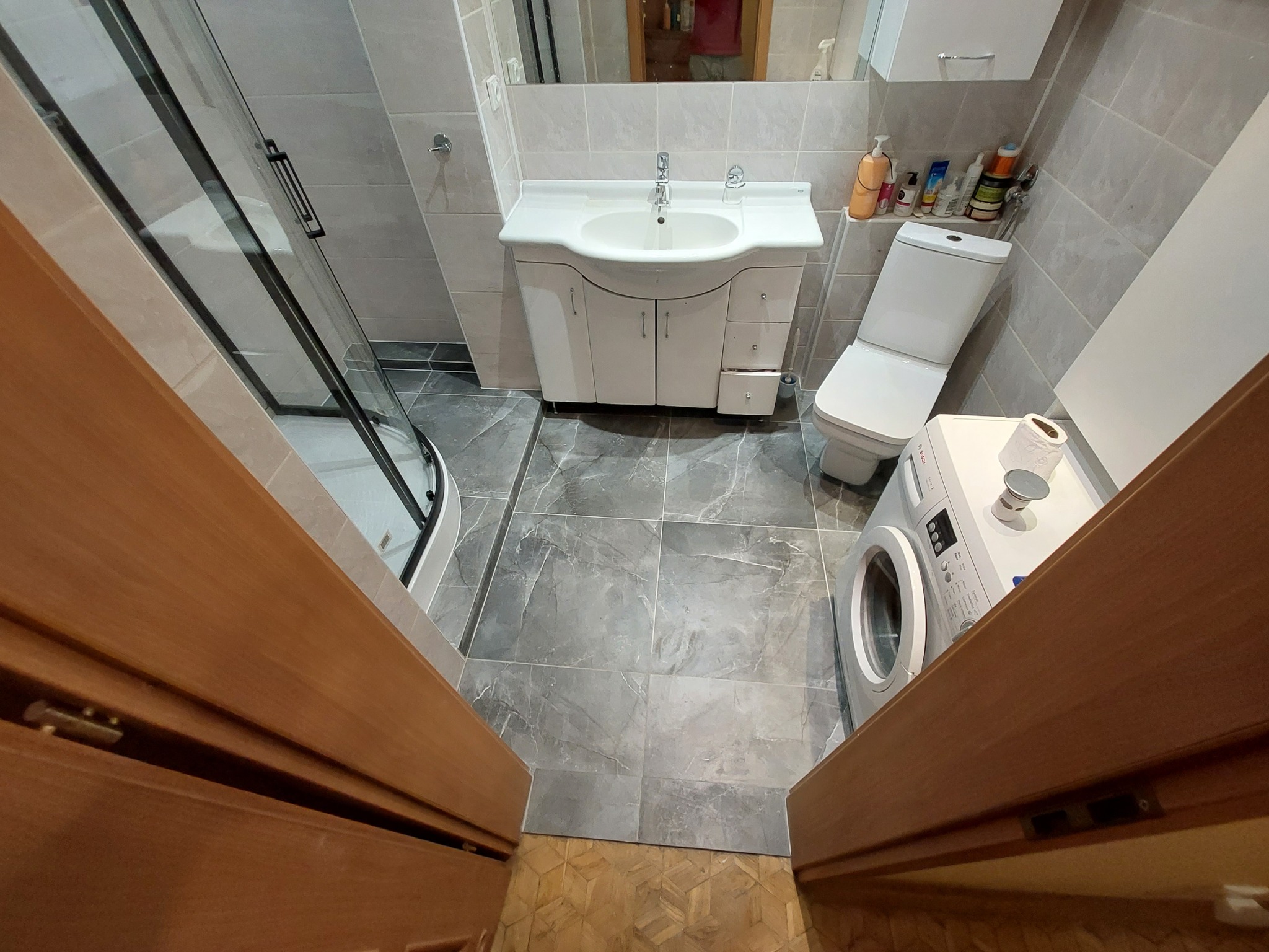 Widok zza progu na wyremontowaną łazienkę z kabiną prysznicową, umywalką z szafką, toaletą i pralką, wyłożoną szarymi płytkami imitującymi marmur.