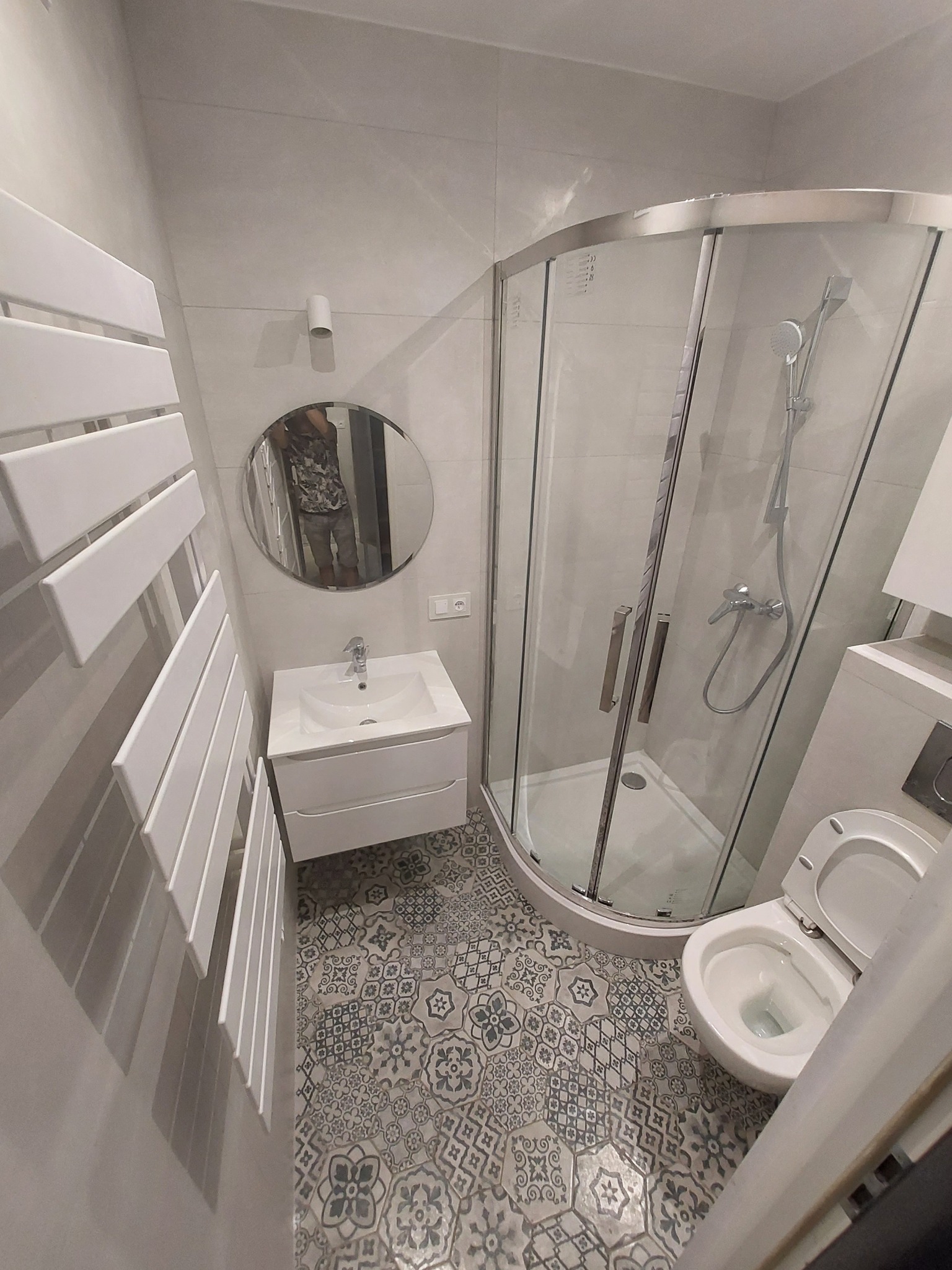 Nowoczesna łazienka z kabiną prysznicową narożną, umywalką z szafką, grzejnikiem drabinkowym i toaletą, wykończona szarymi płytkami i dekoracyjną podłogą w stylu vintage.