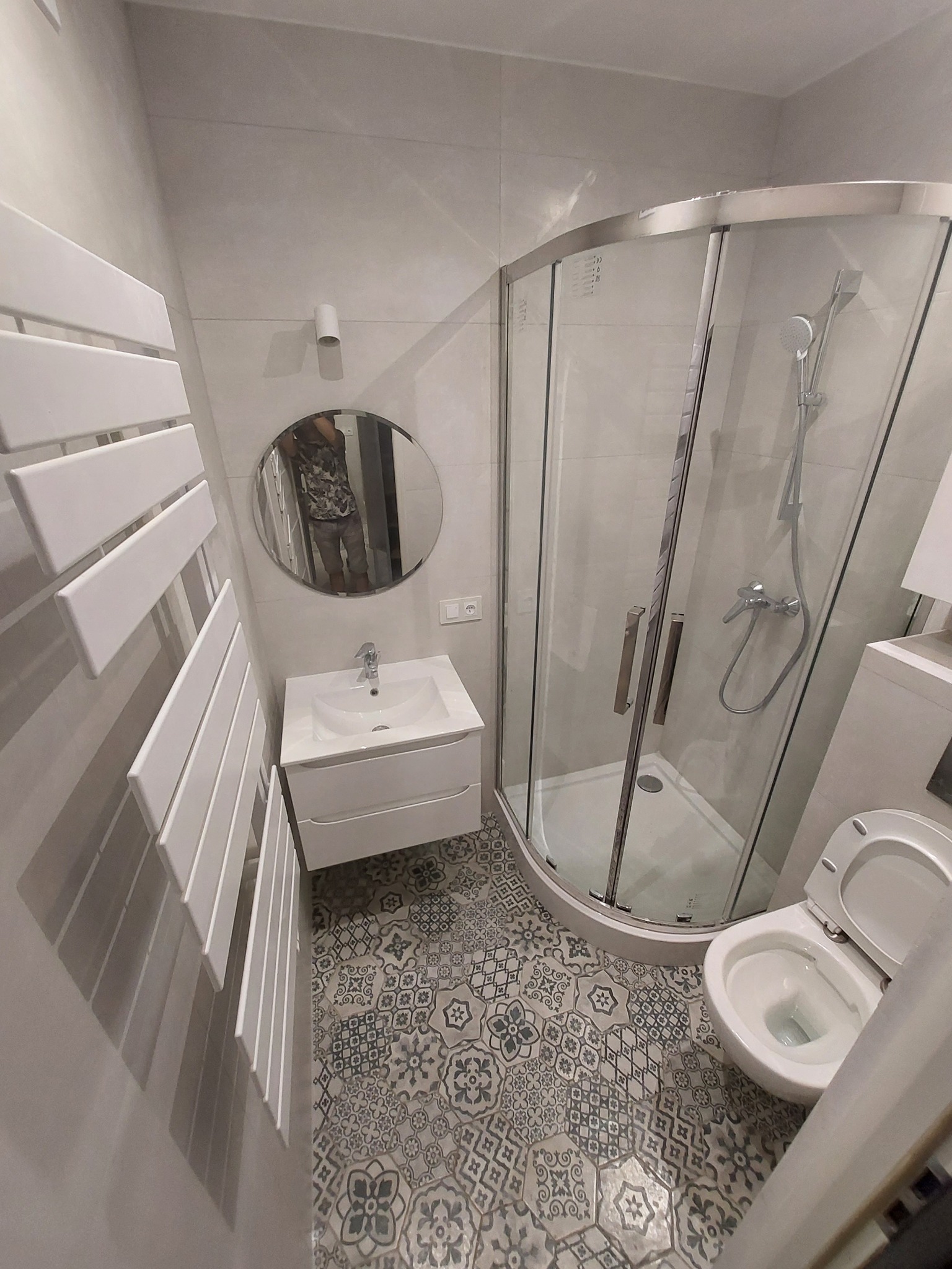 Nowoczesna łazienka z kabiną prysznicową, umywalką z szafką, grzejnikiem drabinkowym i toaletą, wykończona płytkami podłogowymi w stylu patchwork.