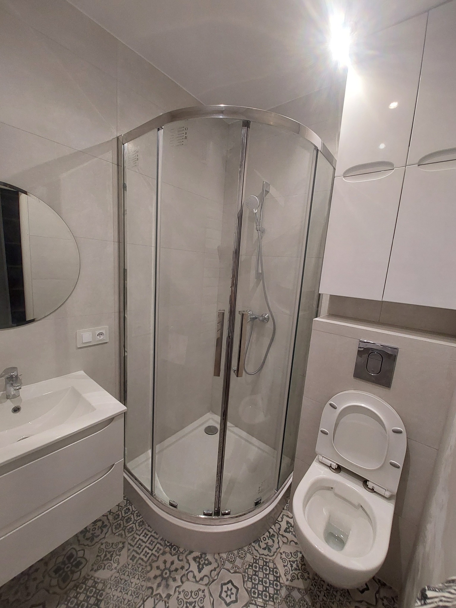 Nowoczesna łazienka z kabiną prysznicową narożną, umywalką z szafką, podwieszaną toaletą i wzorzystą podłogą w odcieniach szarości.