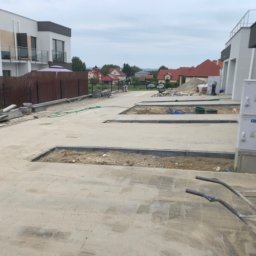 Przejarbruk - Przygotowanie podłoża pod chodnik z kostki brukowej na osiedlu nowych domów jednorodzinnych, widoczne wykopane fragmenty i krawężniki, w tle sterta kostki i taczka.