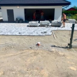 Przejarbruk - Prace brukarskie w toku: częściowo ułożona szara kostka brukowa przed garażem, palety z kostką, pracownicy bez koszulek w tle, widoczny fragment ogrodzenia i przygotowany teren.