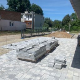 Przejarbruk - Nierówne ułożenie szarej kostki brukowej na piasku, palety z kostką w folii, przygotowany teren pod dalsze prace, widok na domy w tle.