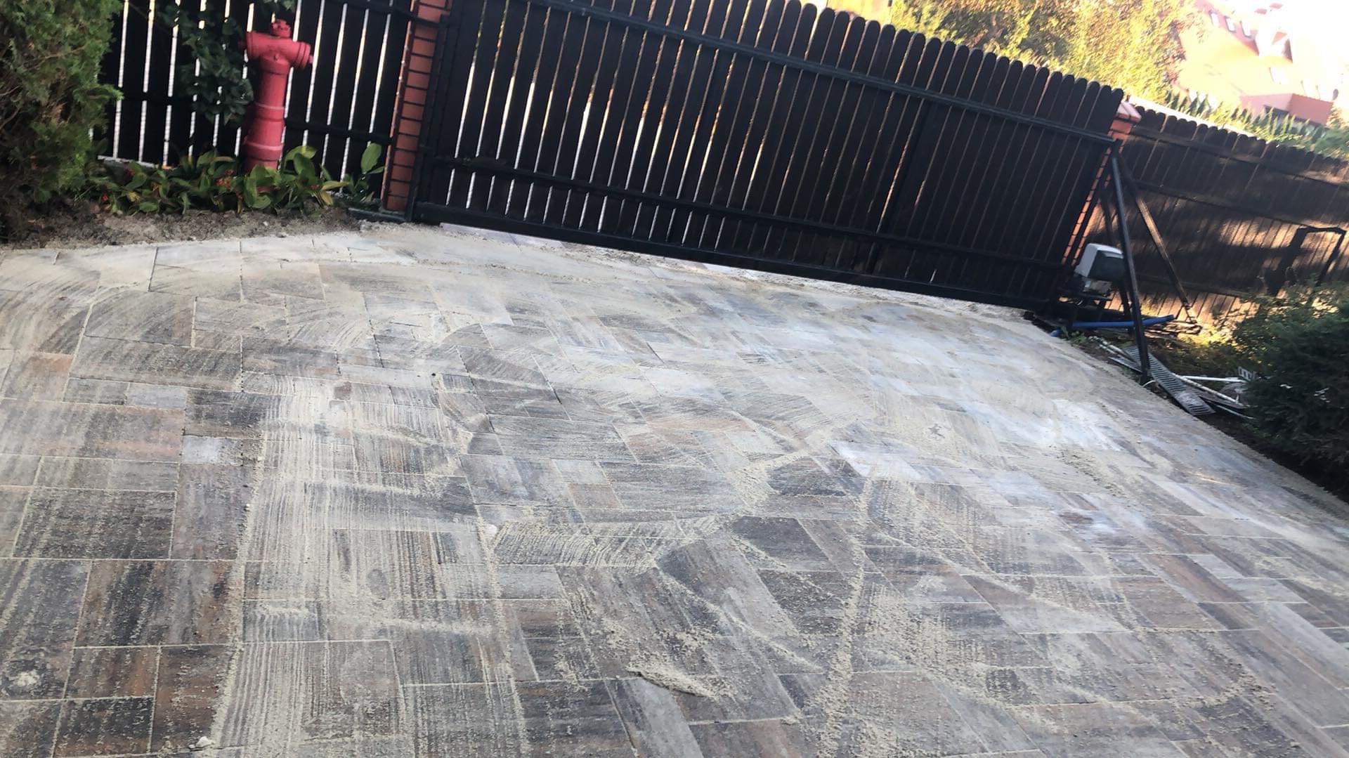 Nowo ułożona, brązowo-szara kostka brukowa z widocznymi śladami piasku, otoczona ciemnym, metalowym ogrodzeniem i zielenią.