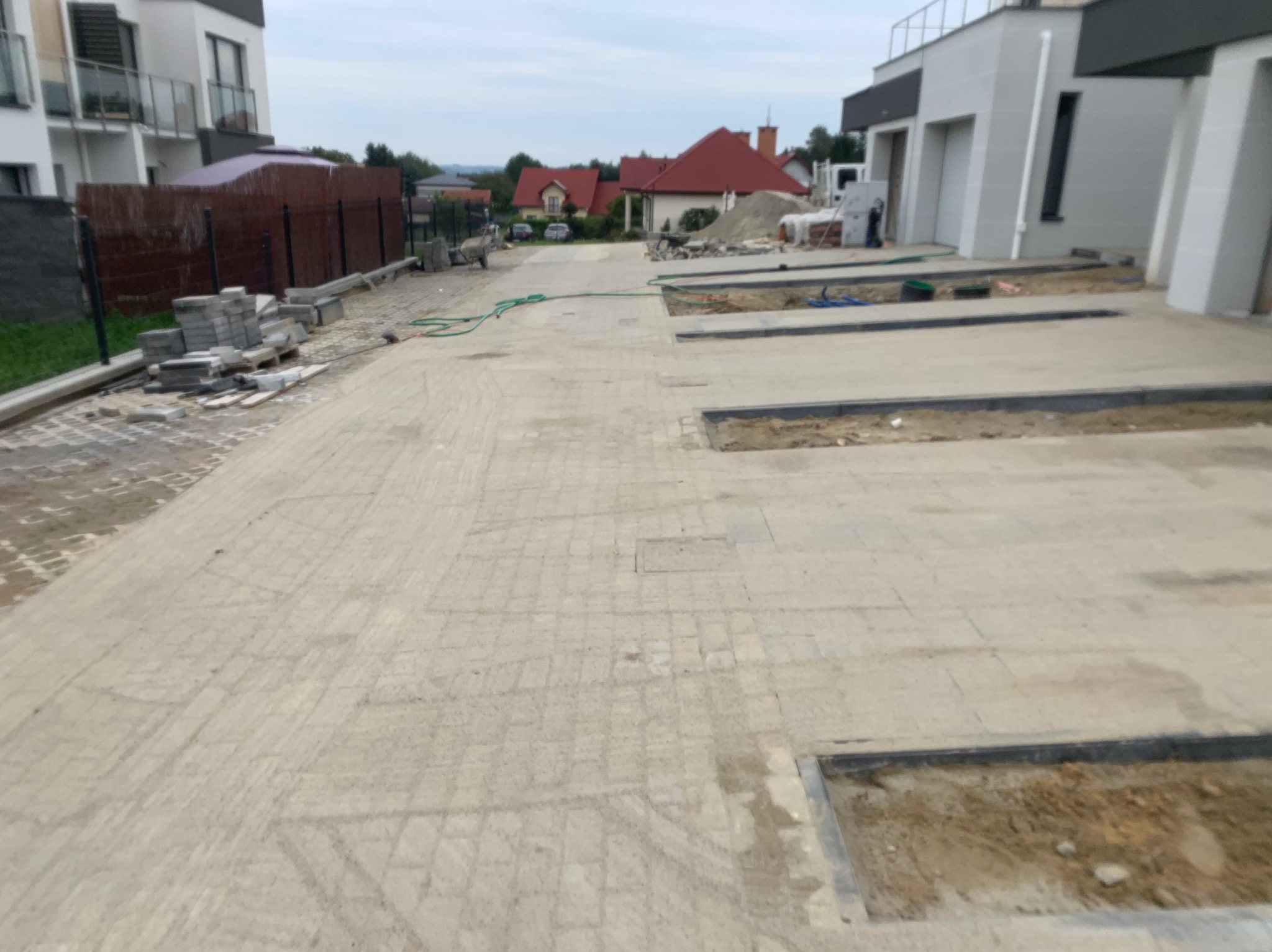 Powierzchnia z jasnej kostki brukowej, częściowo ułożonej, z widocznymi wykopami na obrzeżach, sterty kostki i piasku w tle, oraz domy w zabudowie szeregowej.