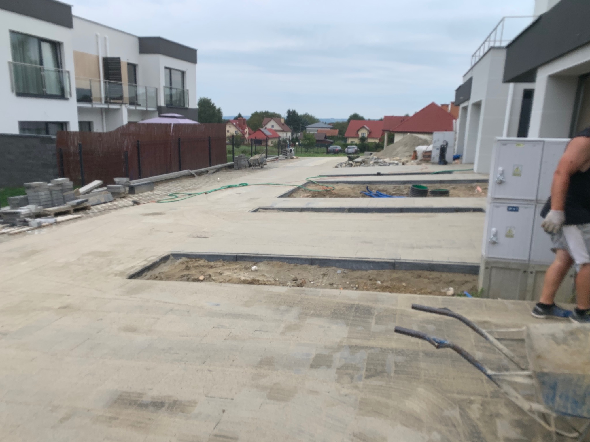 Przygotowanie podłoża pod chodnik z kostki brukowej na osiedlu nowych domów jednorodzinnych, widoczne wykopane fragmenty i krawężniki, w tle sterta kostki i taczka.