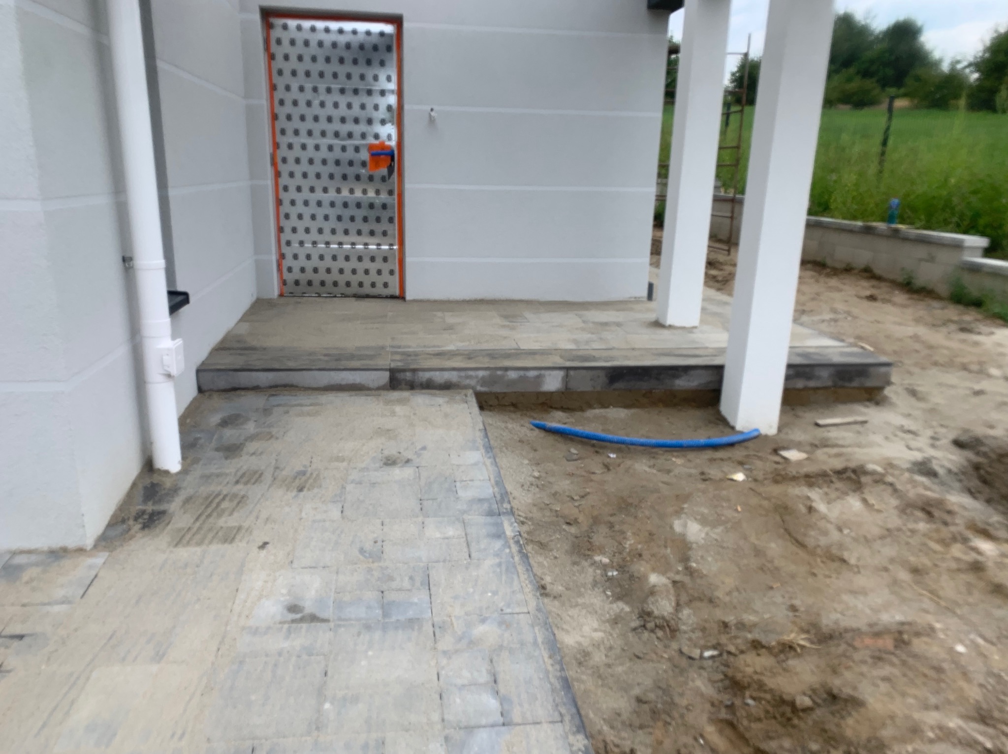 Ułożona szara kostka brukowa prowadząca do wejścia do budynku z charakterystycznymi metalowymi drzwiami z pomarańczową ramą, widoczny fragment wykopu z niebieskim przewodem.