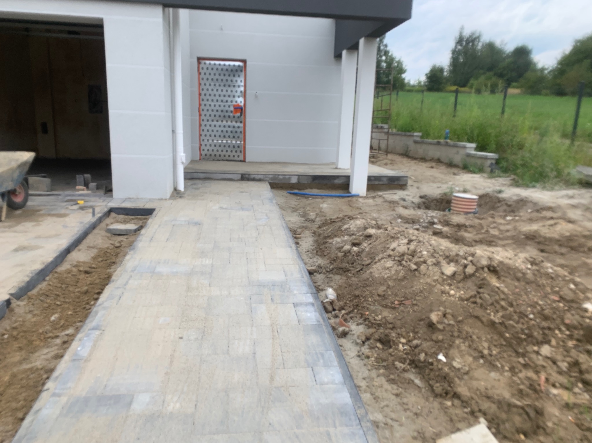 Ułożona kostka brukowa prowadząca do wejścia domu, widoczne przygotowane korytka po bokach i wykop pod przyszłe prace ziemne.
