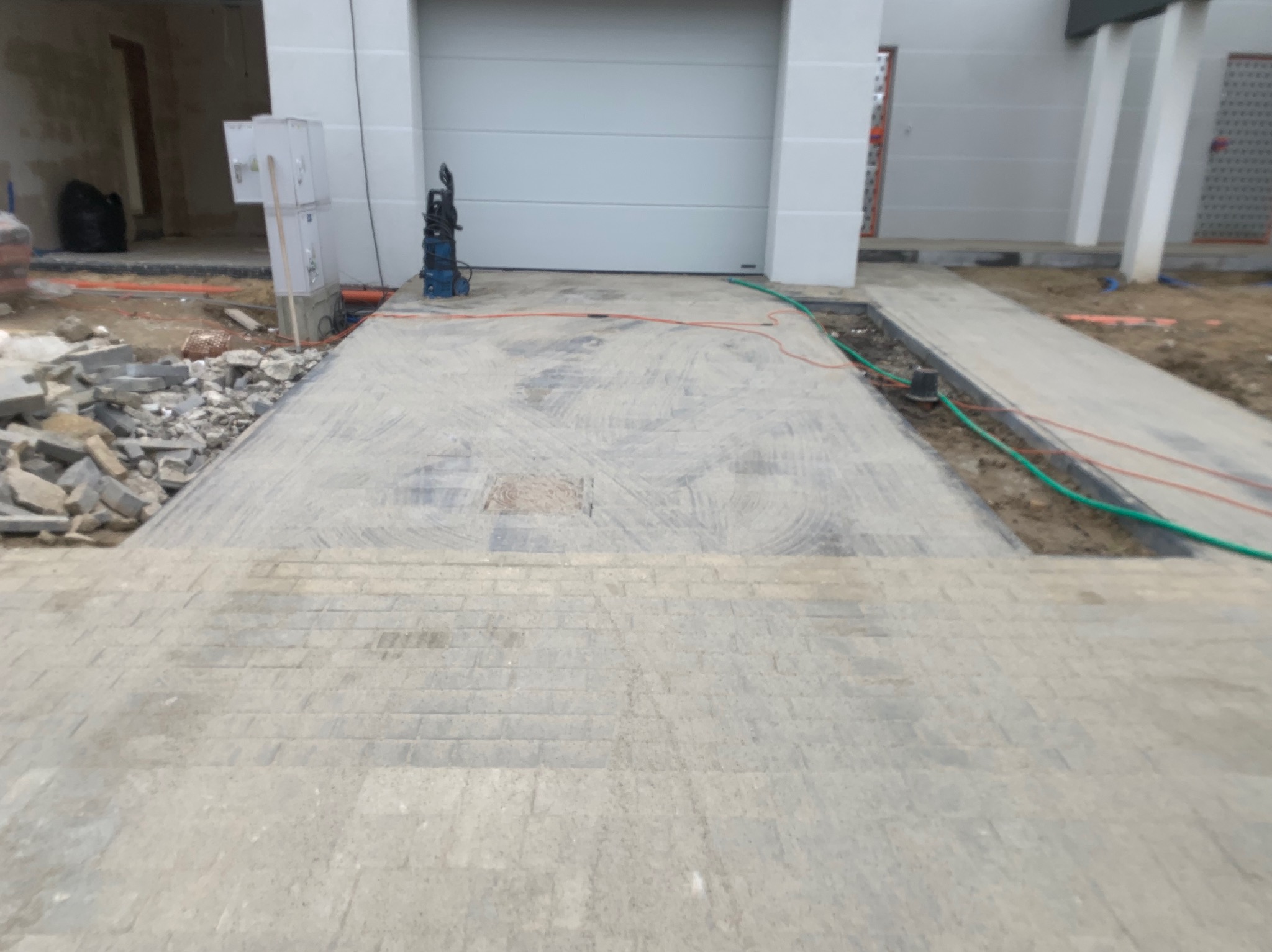 Świeżo ułożona, szara kostka brukowa prowadząca do garażu z widocznymi śladami po czyszczeniu, z resztkami materiałów budowlanych po bokach i myjką ciśnieniową w tle.